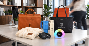 Collection of premium branded corporate gifts displayed on a modern office desk, including a leather briefcase, black tote bag, insulated drinkware, plush blanket, JBL headphones, and illuminated digital clock.