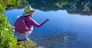 A woman wearing a decorated hat and colorful woven shawl kneels beside a body of water, splashing water with her hand.