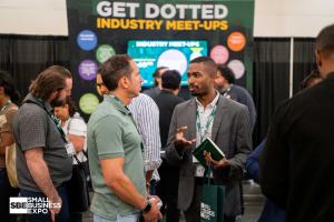 Two attendees speak in front of a Get Dotted Industry Meet-Ups display, with one holding a green notebook and tote bag.