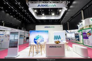 Arkema exhibition booth with suspended white signage, illuminated displays, a reception counter and high-table seating.