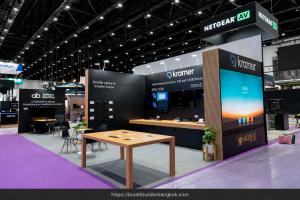 Kramer exhibition stand with black branded walls, a vertical digital screen, demonstration counters and a wooden meeting table.