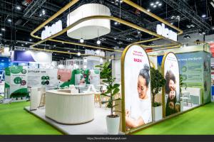 Oleon exhibition booth with illuminated overhead signage, arched portrait panels, a curved white counter and potted plants.