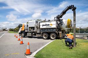 Pulse Locating operator kneeling at a vacuum excavation hole on a grass verge, with the truck's boom lowered and traffic cones along the road