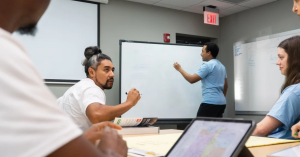 Students in an ACSOM classroom discuss course material around a table while a classmate writes on a whiteboard.