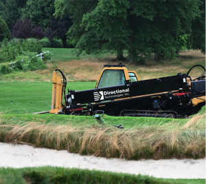 A black and yellow Directional Technologies horizontal drilling rig positioned on grass beside a golf course sand bunker, with mature trees in the background.