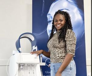 Kierra Fears, founder of Vela Lash Bar, smiles while standing beside a white Glacē treatment device with an attached blue LED arch, in front of a blue Glacē promotional banner.