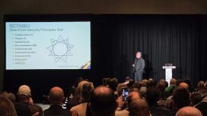 Jim West is standing on a black stage with a lit podium. The stage is flanked by a large screen for his presentation on both sides. On the current slide, his visual Triad is displayed utilizing the nine security principles in cybersecurity.