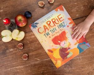 A child’s hand reaches toward Carrie the Tree Fairy: A Fall Fairy Tale, an illustrated hardcover children’s book resting on a table.