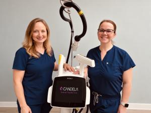 Heather Montgomery, a woman with long blonde hair, and Sarah Scaggs, a woman with brown hair and glasses, both wearing navy blue scrubs, stand smiling on either side of a white Candela Nordlys laser and IPL system, whose screen displays the Candela logo a