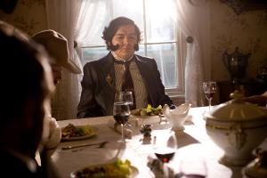 Production still from Interrupted Passage by Julio César Morales and Max La Rivière-Hedrick: a costumed actor portraying Gen. Mariano Guadalupe Vallejo sits at a candlelit dinner table set with wine glasses and plated food, with guests in the foreground.