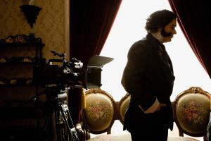 Production still from Interrupted Passage by Julio César Morales and Max La Rivière-Hedrick: an actor in 1840s costume as Gen. Mariano Guadalupe Vallejo stands in silhouette by a sunlit window beside a film camera and antique upholstered chairs.
