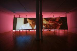 Installation view of Interrupted Passage by Julio César Morales and Max La Rivière-Hedrick: two large video screens on stands in a pink-lit gallery, one showing a person in a white shirt looking down, the other a close-up of food being prepared.