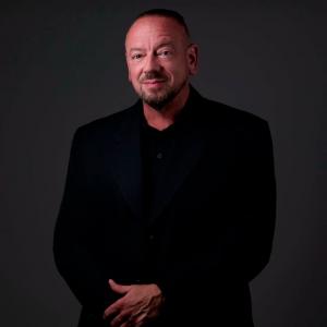 Author David M. Ward wearing a black suit and black shirt against a dark gray background.