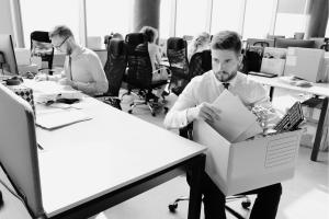 A young man looking dejected who was recently part of a layoff, sitting at his former workspace with co-workers behind him and his personal belongings in lap.
