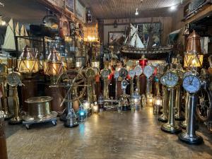 Display of brass engine order telegraphs, illuminated copper maritime lanterns, ship's wheels and a large model sailing ship inside a nautical antiques showroom.