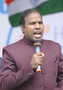 Dr. K. Paul speaking into a microphone while wearing a maroon formal jacket, with a partially visible Indian flag in the background.
