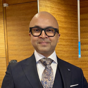Ratib Zaman wearing a dark pinstripe suit, patterned tie and black glasses, standing in front of a wooden wall.