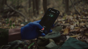 A gloved hand holds a smartphone running a Deep Breathe pneumothorax ultrasound scan, outdoors on a forest floor.