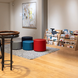 Cozy reading nook with colorful stools, a magazine rack, staircase, round wooden table, and abstract wall art.