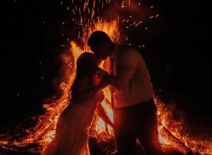 A bride and groom share a kiss in front of a blazing bonfire at night, surrounded by glowing flames and flying sparks, creating a romantic, rustic wedding atmosphere.