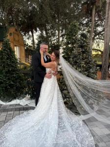 Bride and groom embrace in an outdoor wedding portrait surrounded by decorated Christmas trees with twinkling lights, a rustic wooden barn, and a long, flowing bridal veil.