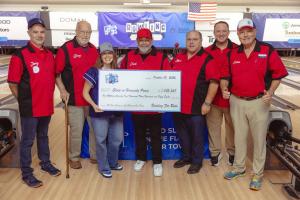 Seven people in red-and-black bowling shirts and SHP caps stand on a bowling lane holding a giant check to Sleep in Heavenly Peace for $1,072,358.