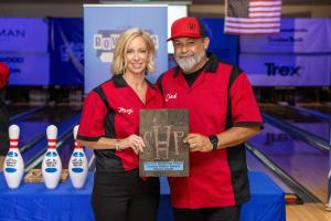 Margi Vagell and Chad Sanders, in matching red-and-black bowling shirts, hold a wooden plaque with a copper SHP logo engraved “Dream Maker Award, Margi Vagell.”