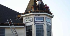 Roofers working on a shingled turret roof above bay windows, with a ladder against the house