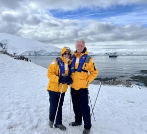 Chip Walter and Cyndy Mosites stand among penguins at the short of D'Hainaut Island, Antarctica. Snow covered islands and ocean fill the background.