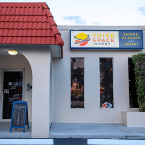 Exterior of Third Space Improv comedy theater with red tile roof, yellow-and-blue sign, entrance, and sidewalk sandwich board.