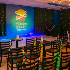 Colorful comedy theater seating decorated with leis, facing a stage and “Third Space Improv” sign beneath starry curtains.