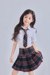 Seven-year-old Australian-Indonesian young girl Arrow Travnick looking off to the side, wearing a white short-sleeved collared shirt, a red-and-green plaid tie and a matching plaid pleated skirt against a plain light gray backdrop.