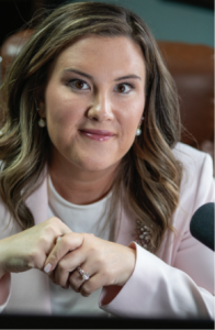 Whitney Lundy Pennington smiling at the camera, with long, wavy, highlighted brown hair and pearl drop earrings. She wears a pale pink blazer over a white top, hands clasped in front of her beside a microphone.