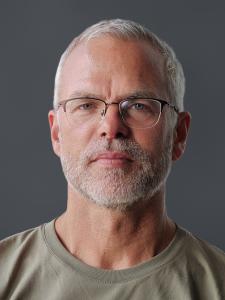 Aris Hägermann mit Brille und olivfarbenem T-Shirt vor grauem Hintergrund