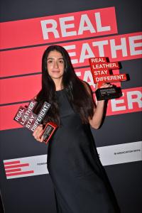 Israeli fashion designer Tal Zohar smiles while holding two Real Leather. Stay Different. 2026 competition trophies against a red and black branded backdrop at the Bangkok international final.