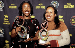 Locsanity Community Impact Award winners Ijeoma Anyanwu (left) and Monique Nicholas (right) smile while holding colorful glass trophies in front of a LOCnificent 2026 event backdrop.