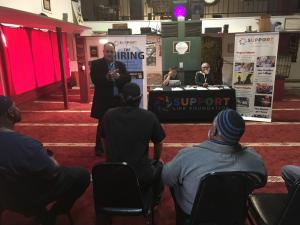 A man addresses a diverse crowd seated in front of a Support Life booth