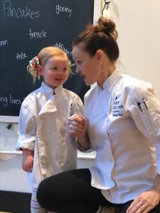 Founder of LKA kneeling down wearing chef coat next to young girl student in chef coat