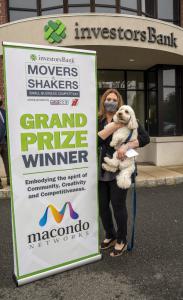 Jennifer Frey, managing partner of Macondo Networks, is holding Dash, a golden doodle dog, which is the company mascot. Frey is standing next a sign that Grand Prize winner, Movers & Shakers competition.