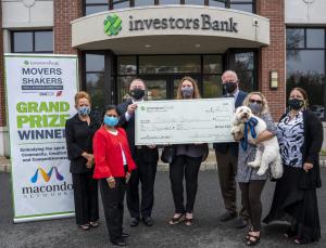Seven people standing in front of an Investors Bank branch. Three people are holding a large check. On the left side is Movers & Shakers Small Busness Competition sign. On the far right a woman is hold a golden doodle dog, named Dash.