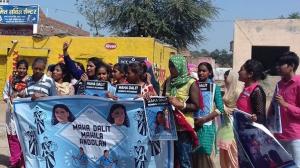 A group of women holding a blue banner, protest on the street to call for greater rights and protections for Dalit women and girls; it is daytime and the sky in the background is blue