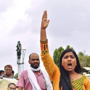 Manisha Mashaal, founder of Swabhiman Society at a street demonstration. She is holding her left arm straight up, pointing at the blue sky, and her mouth is open as if she is speaking. In the background walking behind her are two men taking part in the de
