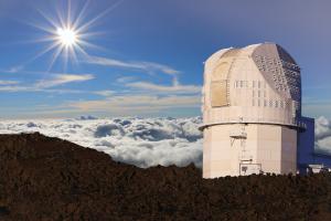 The Inouye Solar Telescope stands tall above the clouds, on the brown-red rocks of Haleakalā. The near violet sky contrasts the bright, beaming sun in the sky above the telescope.