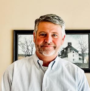 man standing in front of a photo with a gray striped shirt and gray hair and beard