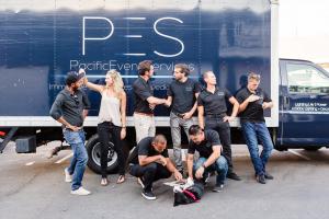 A group of people laughing with a large truck beind them. The truck is black and says Pacific Event Services in white letters