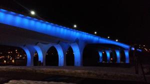 Photograph of the underside of the 35W Bridge over the Mississippi River in Minneapolis. The Bridge is illuminated in blue lights.
