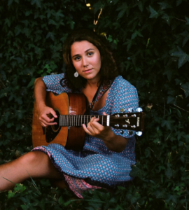 Portland based singer/songwriter Olivia Klugman sits in green ivy holding a guitar