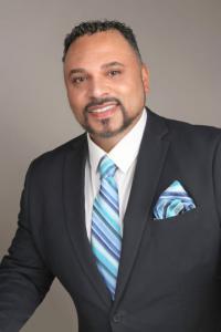 Mike Dunston is in a posed headshot in a dark colored suit, white-collared shirt, with a blue tie and matching blue handkerchief.