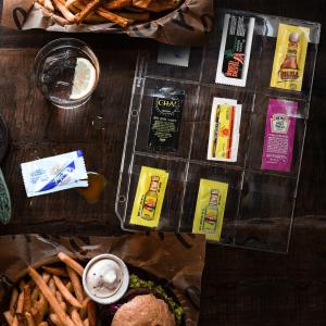 A restaurant table with hamburgers and a binder page filled with hot sauce packets.