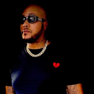 Bigg Brass dressed in black T-Shirt with black sunglasses and thick gold chain around his neck.
